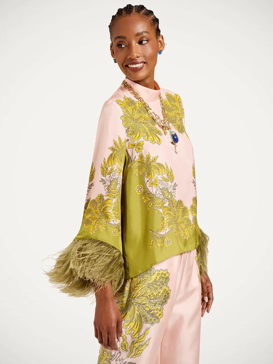 A model wearing a light pink satin poncho-style top with olive green and yellow botanical patterns and feathered cuffs, paired with matching pink trousers.